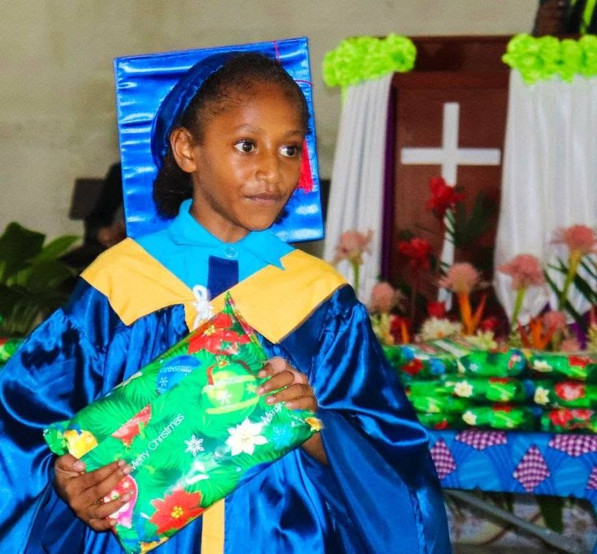 A student in a graduation gown and cap holding a wrapped gift at a PMCA graduation ceremony