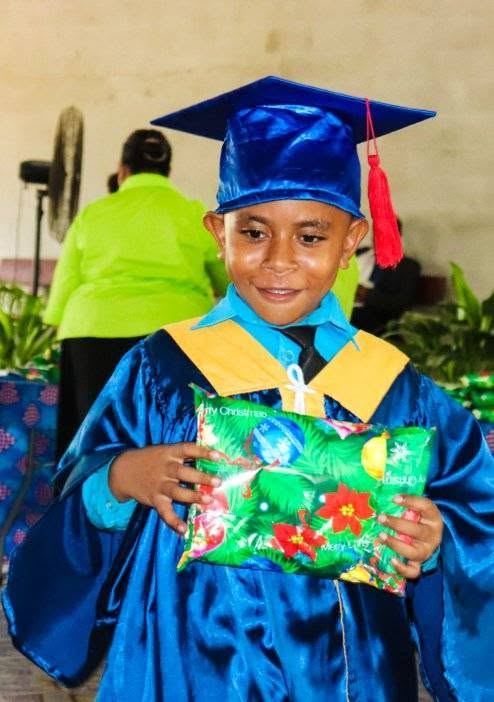 A student in a graduation cap and gown holding a wrapped gift, smiling at a PMCA graduation ceremony